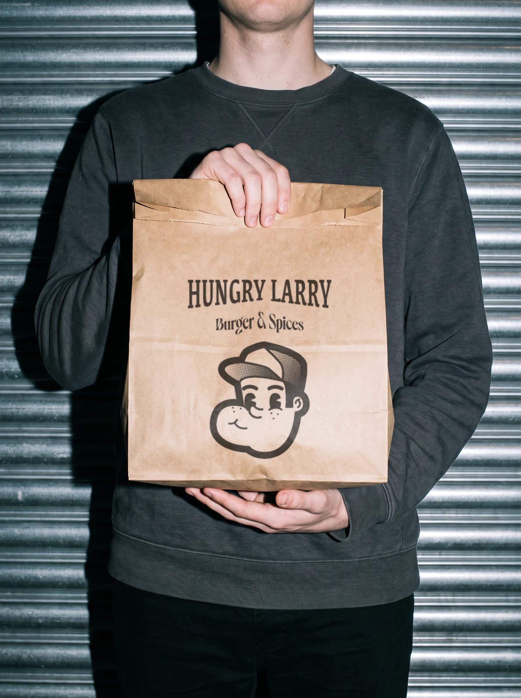 Sac kraft Hungry Larry tenu à deux mains devant un rideau de fer, flash direct